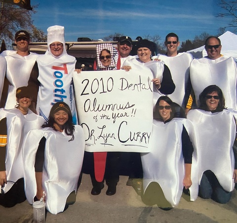 Dr. Lynn Curry smiles for a picture with firends dressed as teeth, toothpaste, and tooth brush. They are holding a sign that says, "2010 Dental Alumnus of the year, Dr. Lynn Curry."