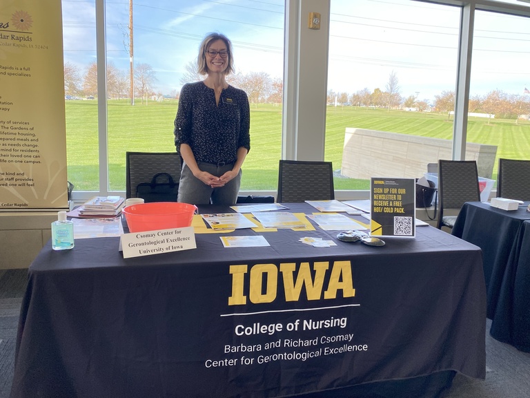 A person in a black shirt stands behind a tablecloth with a logo that reads, "Iowa, College of Nursing, Barbara and Richard Csomay Center for Gerontological Excellence."
