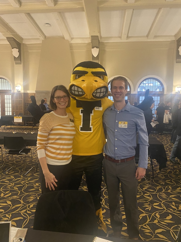 Two people are standing and smiling in a ballroom. Herky, the Hawkeyes mascot, stands between them.