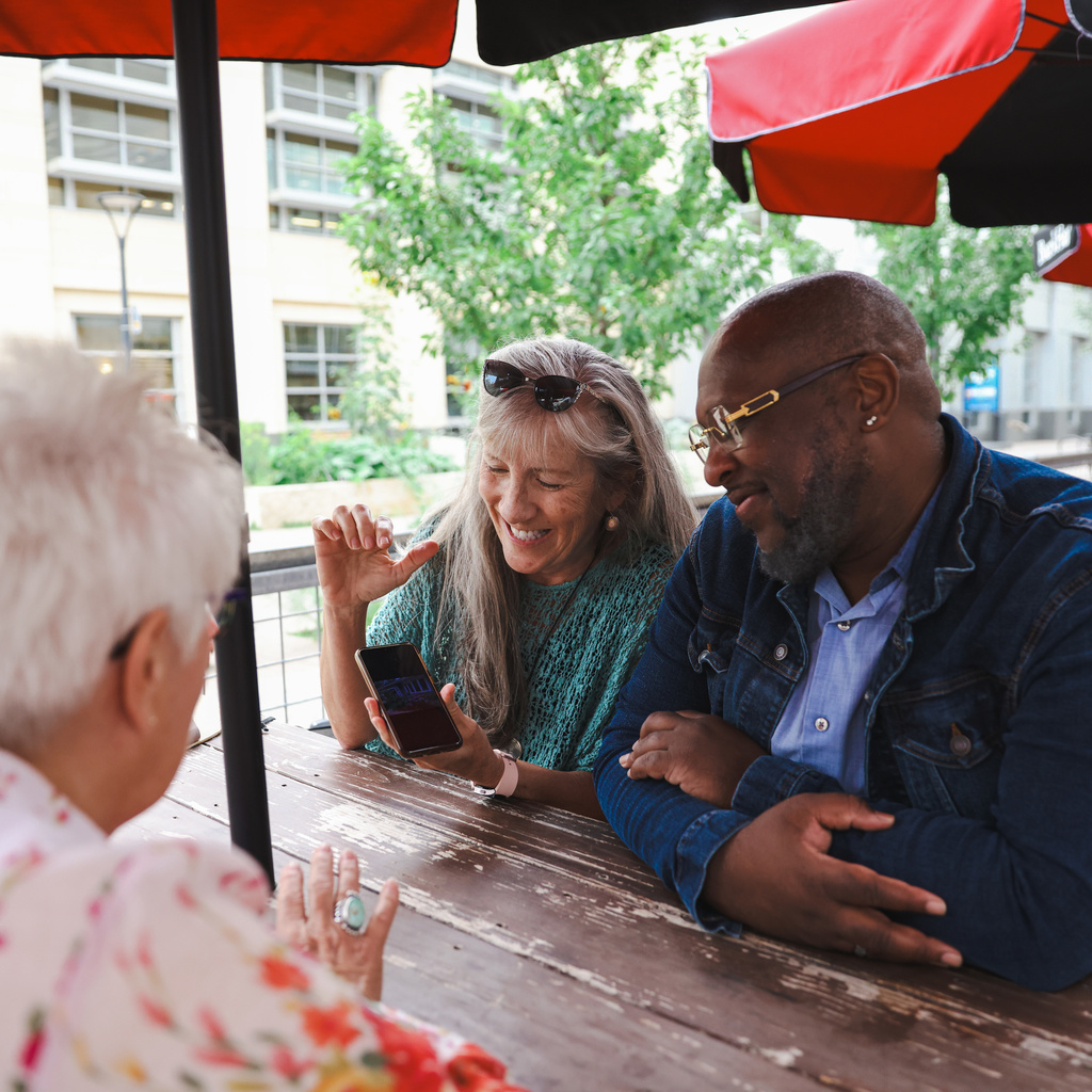 Three people sit around a table under an umbrella. Two have grey hair. They are smiling and looking at a cell phone.