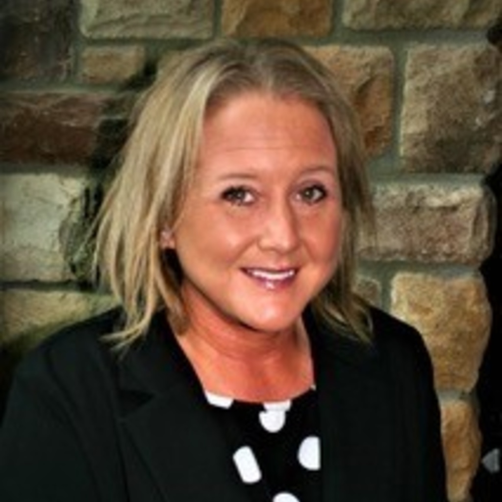 A blond woman smiles in front of a brick wall. She is wearing a shirt with black and white polka dots under a black jacket.