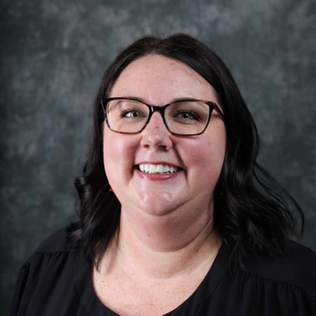 A woman wearing a black shirt and glasses smiles in front of a gray backdrop.