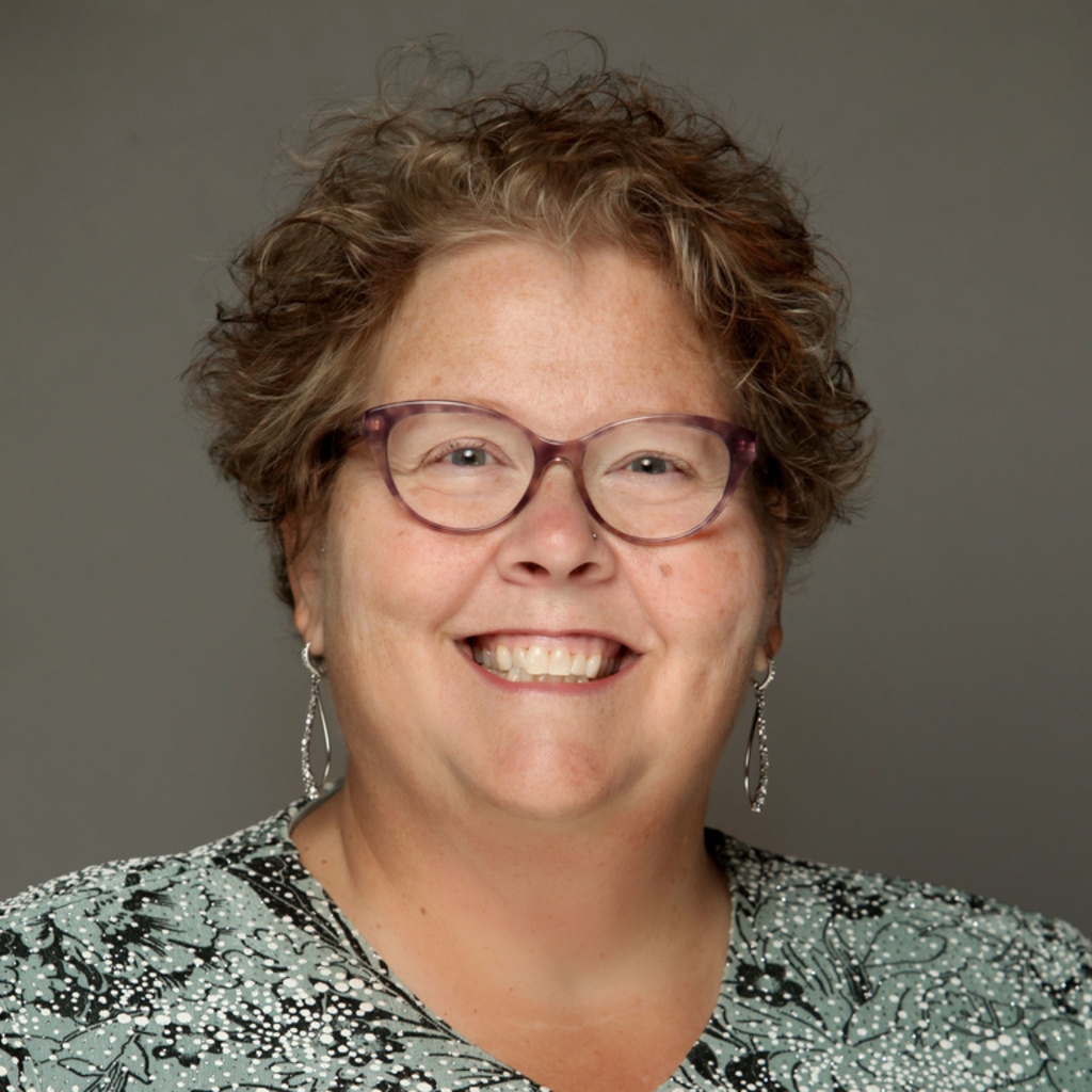A woman with curly hair, glasses, and a light green shirt smiles for the camera.