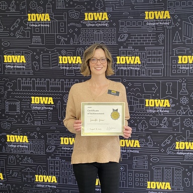 A woman with short hair and glasses stands in front of a banner holding a certificate of completion.