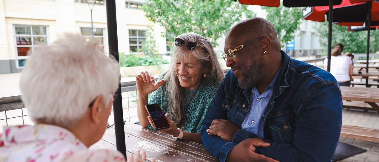 Three people sit around a table under an umbrella. Two have grey hair. They are smiling and looking at a cell phone.