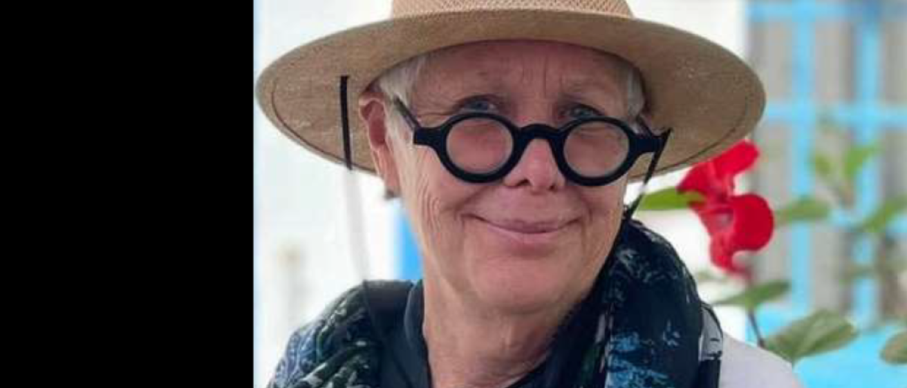 A woman with round black glasses, a straw hat, and a navy scarf smiles at the camera. 