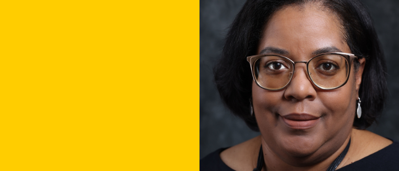 A woman with glasses and short black hair smiles in front of a gray backdrop.