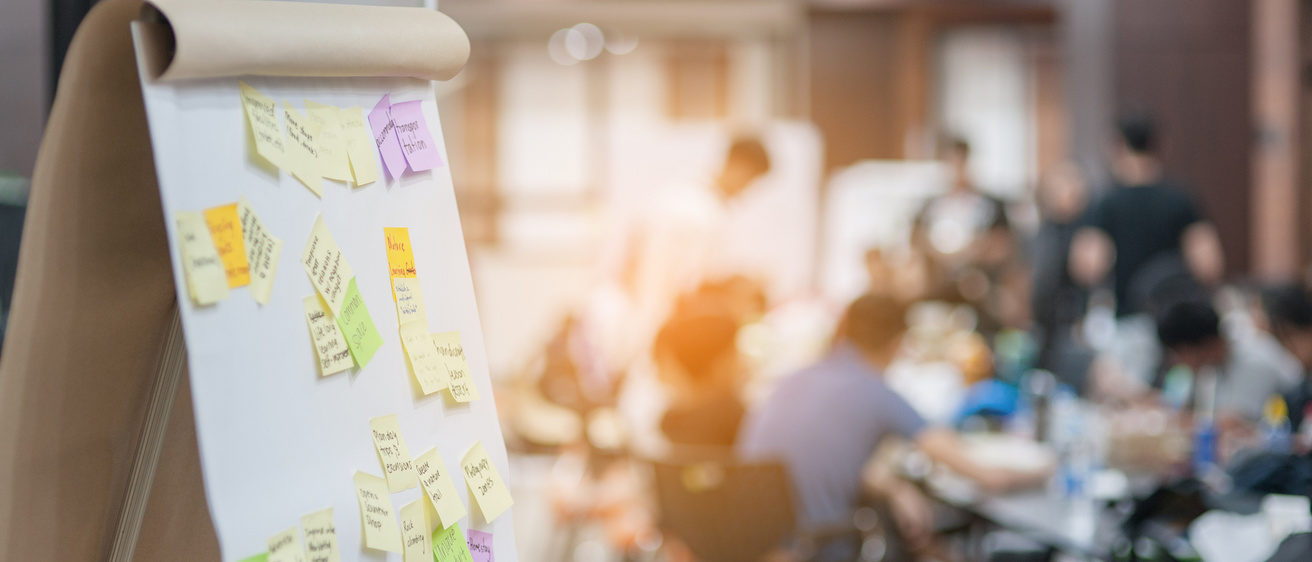 A pad of chart paper covered in brightly covered sticky notes in the foreground, a group of people meet around a table in the background.
