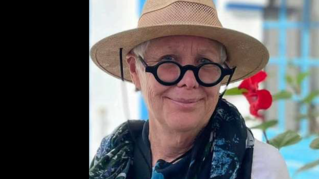 A woman with round black glasses, a straw hat, and a navy scarf smiles at the camera. 
