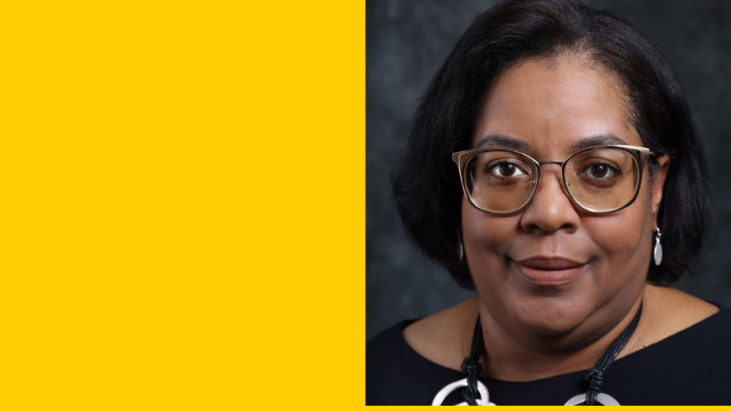 A woman with glasses and short black hair smiles in front of a gray backdrop.