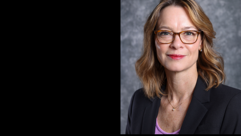 A woman with square glasses, short blond hair, wearing a purple top and black blazer smiles in front of a gray backdrop.